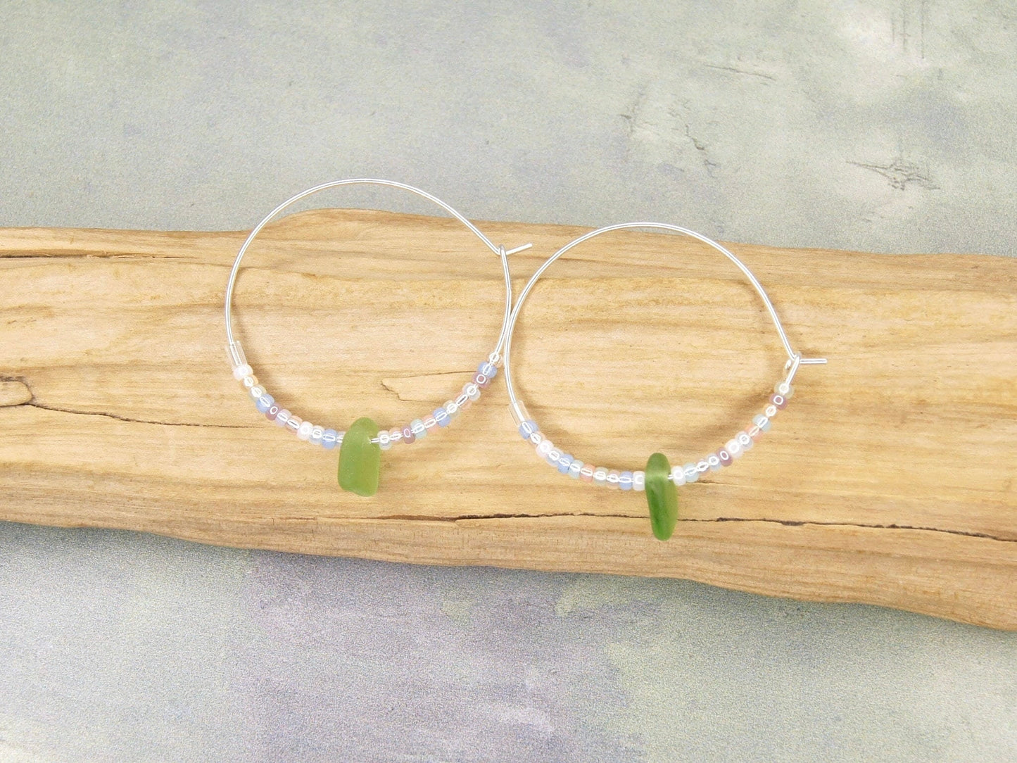 Silver hoop earrings with pastel seed beads and green sea glass laid on a piece of driftwood.