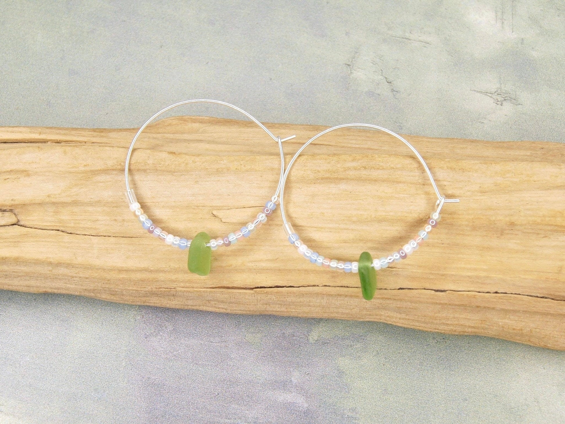 Silver hoop earrings with pastel seed beads and green sea glass laid on a piece of driftwood.