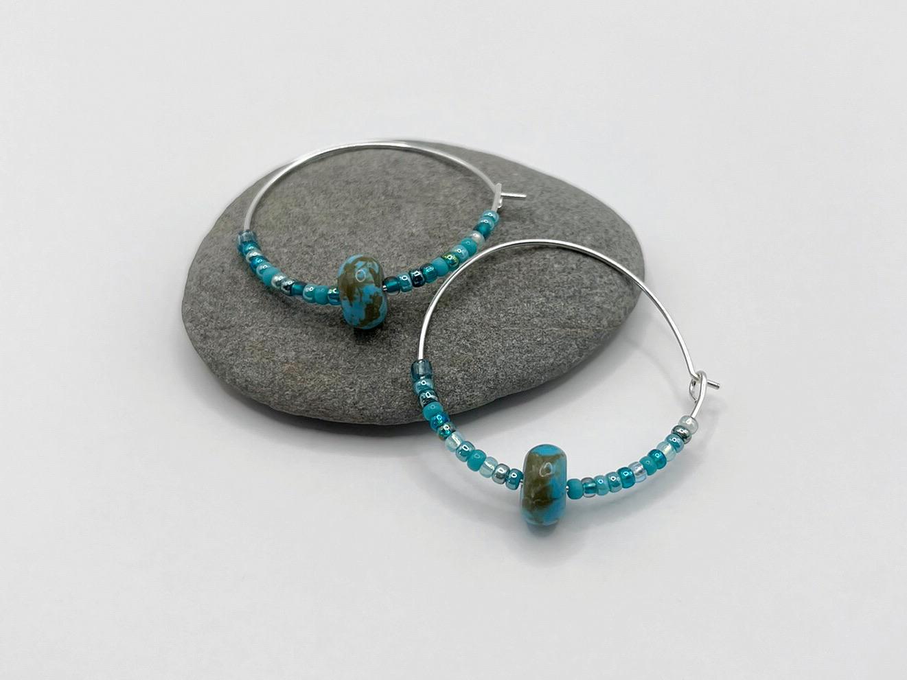 Pair of turquoise beaded hoop earrings laid on a grey pebble.