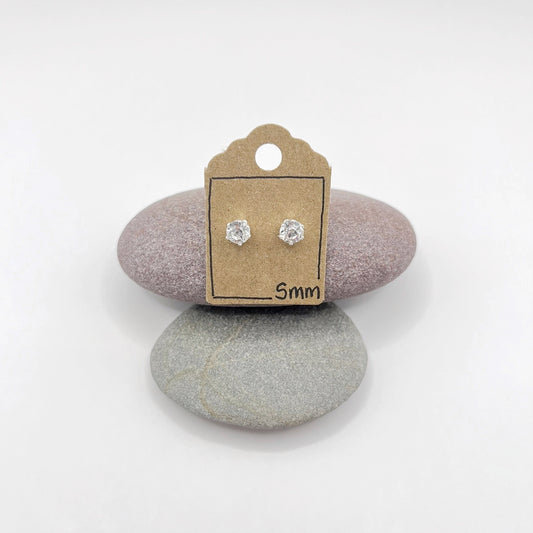 Pair of silver stud earrings with clear cut glass on a small cardboard stand with '5mm' label, placed on two stones.