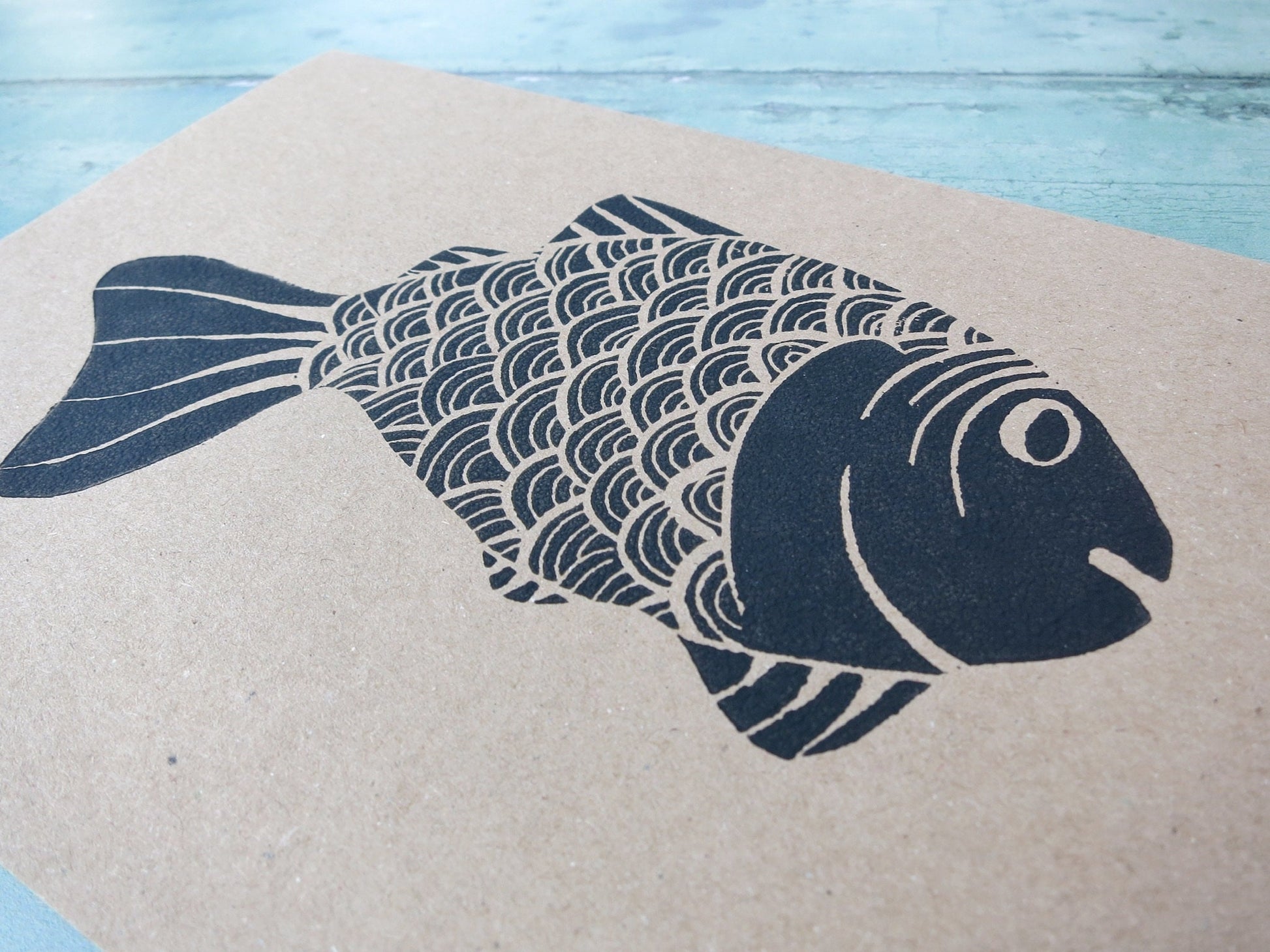close up of Kraft paper card with original lino printed fish laid on a textured surface. 