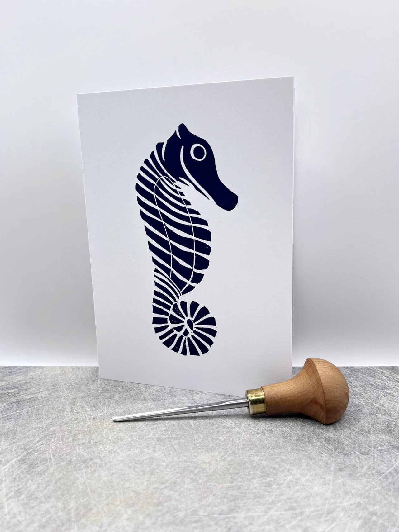 Original lino print of a navy blue seahorse design on a white card, stood against a white background. Photo includes lino cutting tool.
