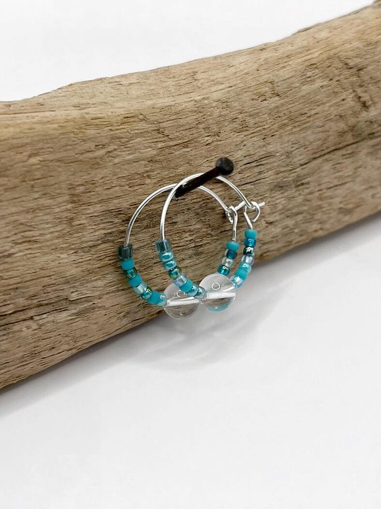 Silver hoop earrings with turquoise seed beads and crystal hanging on driftwood against a white background