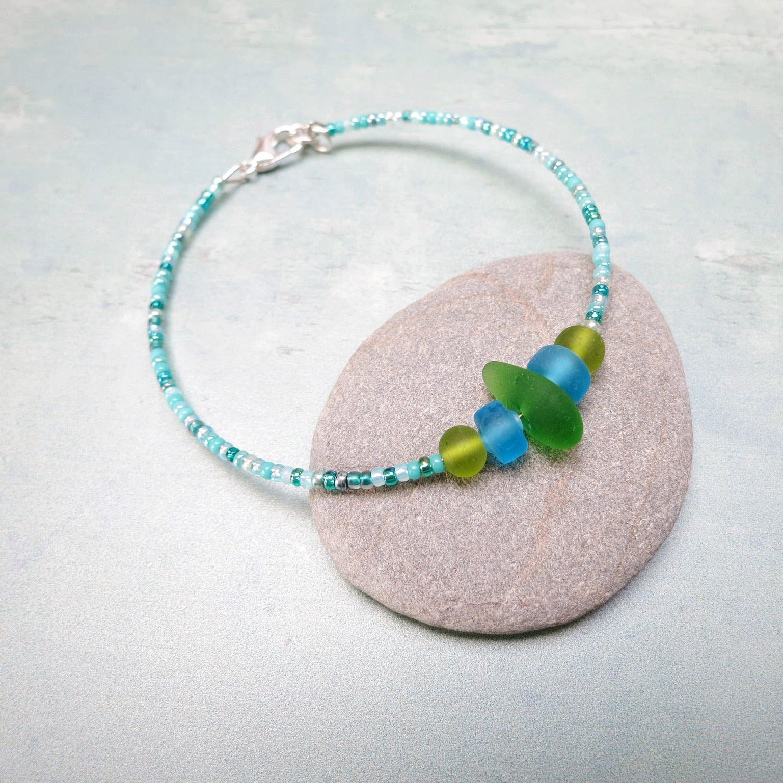 Bracelet with green sea glass and turquoise and green glass beads laid on a purple pebble against a pale blue background