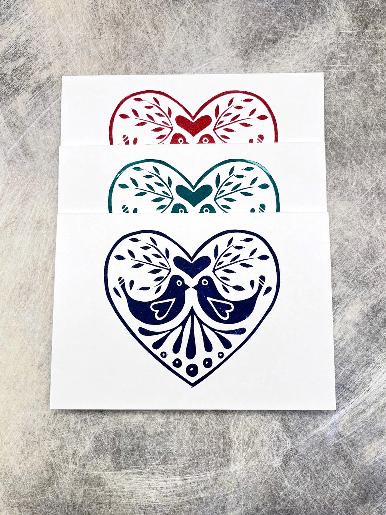 Set of three original lino printed heart design cards in red, blue and green, laid on a  grey textured background.
