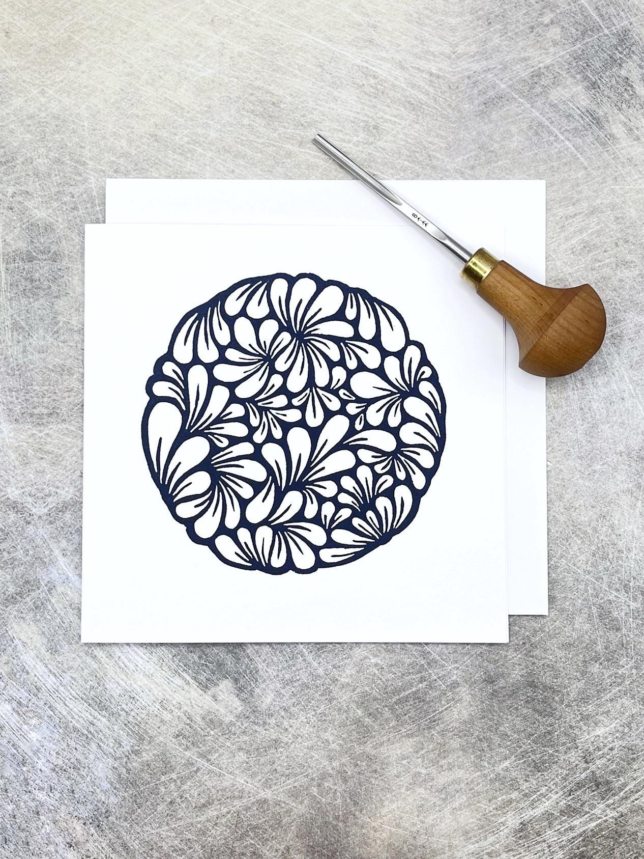 Original navy blue lino printed mussel shell pattern on white card laid on a textured background. Photo includes lino cutting tool.