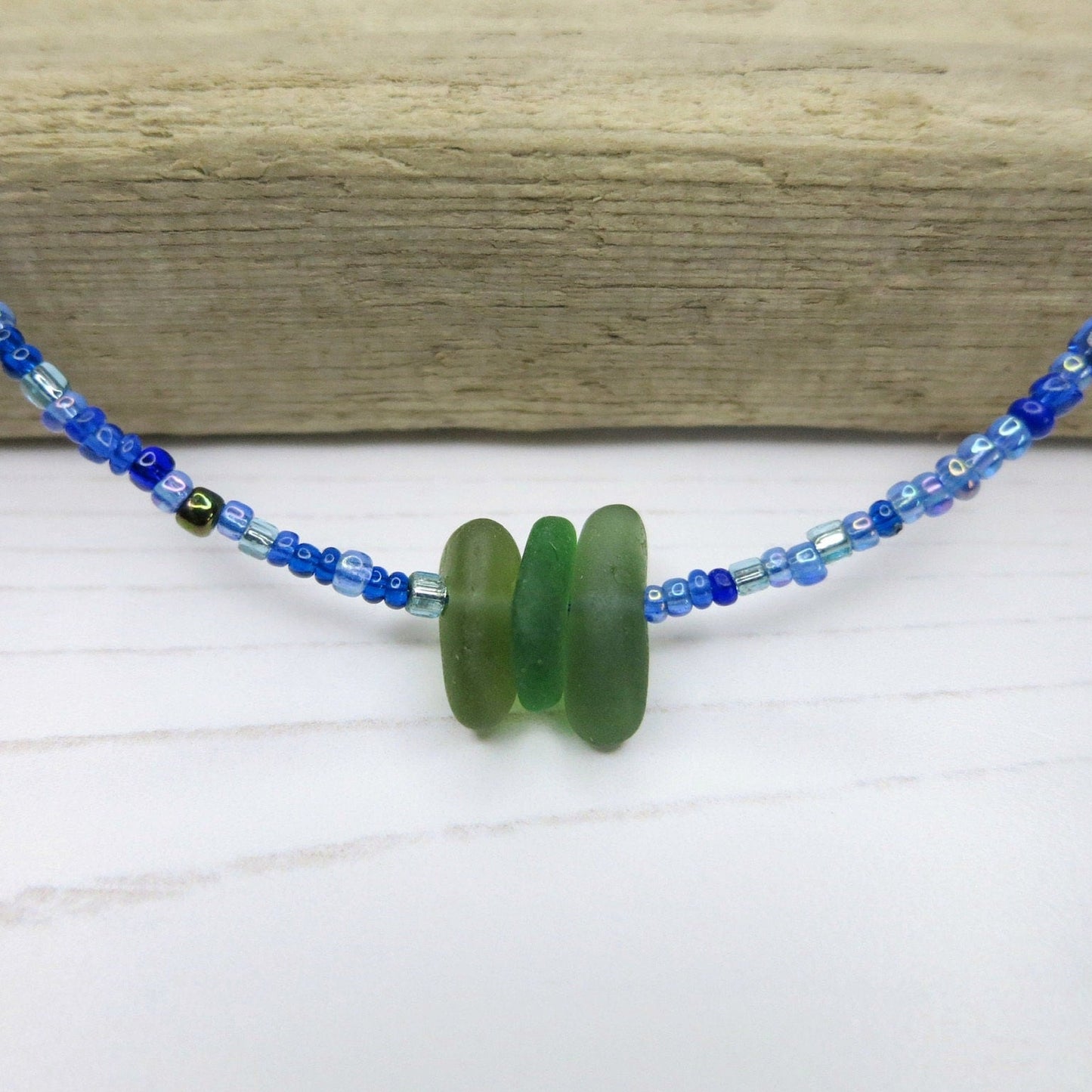 Close up of blue seed bead necklace with green sea glass on a white surface