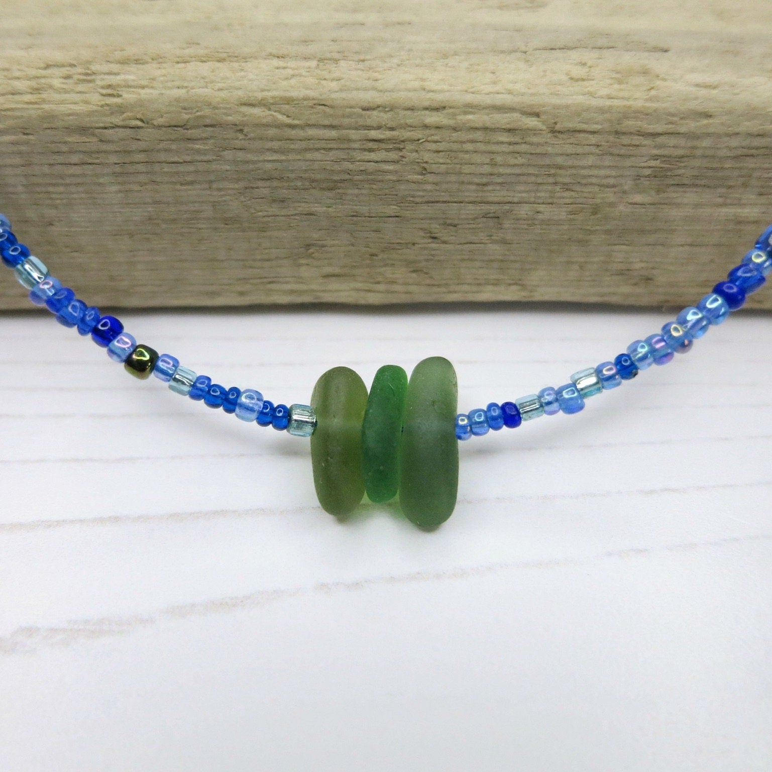 Close up of blue seed bead necklace with green sea glass on a white surface