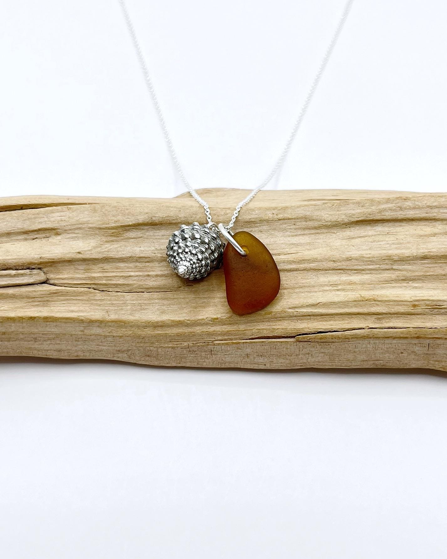 Necklace with burnt orange sea glass and silver shell pendant laid on a driftwood against a white background