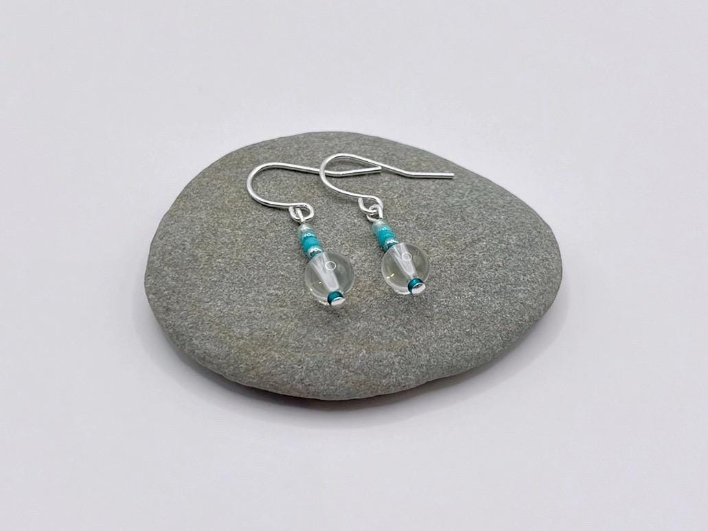 Silver earrings with clear crystal and turquoise seed beads laid on a grey pebble.