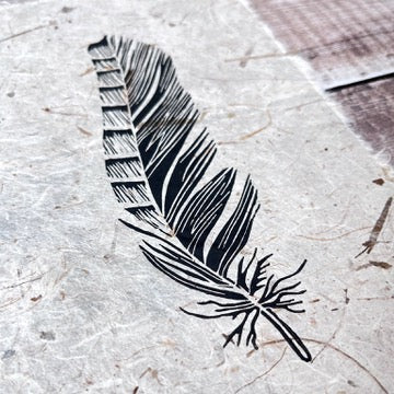 Close up of lino print of a Jay feather in black ink on handmade paper. Laid on a wooden background.