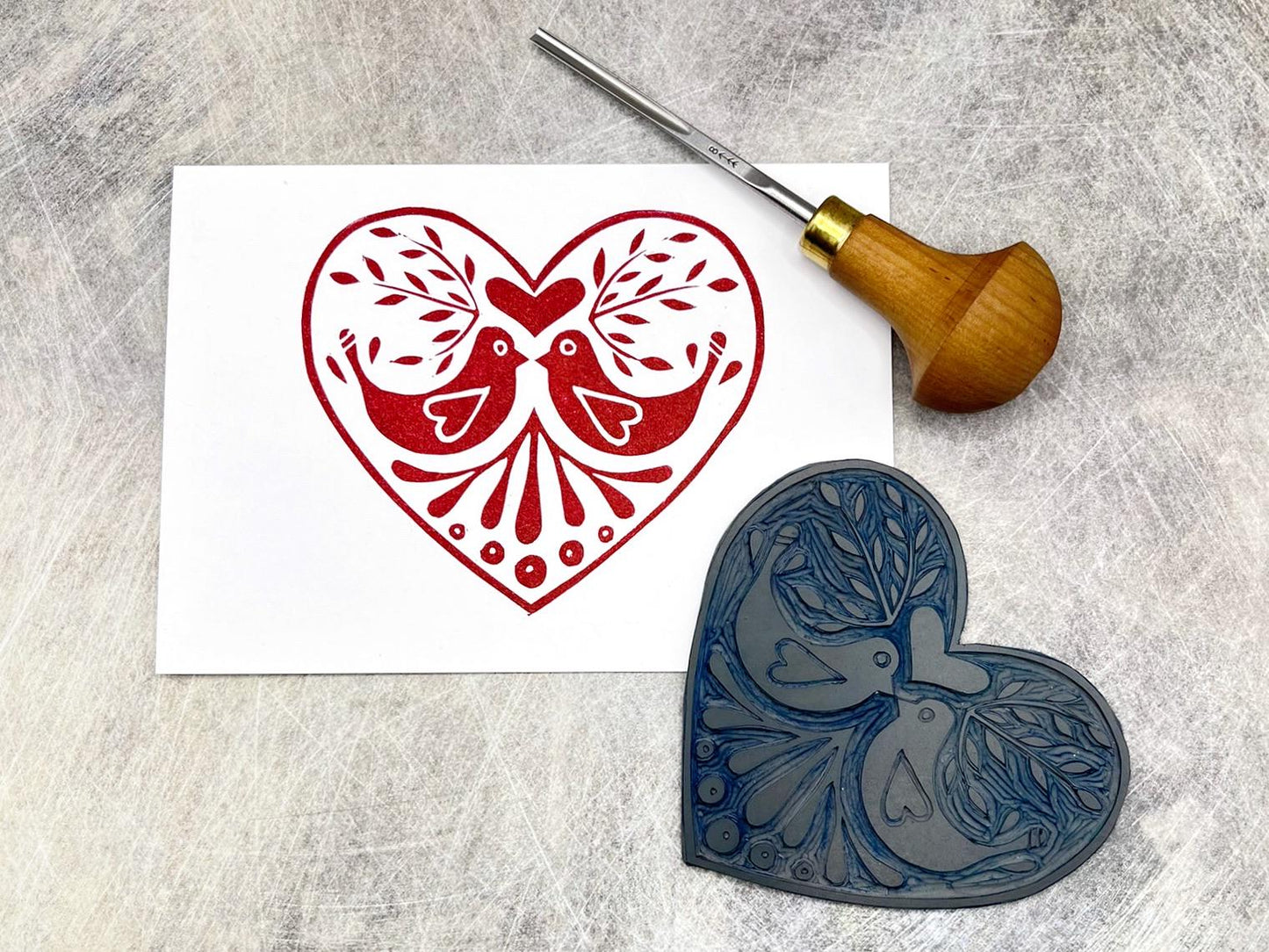White card with original lino printed red heart design featuring two love birds. Photo includes lino and cutting tool.