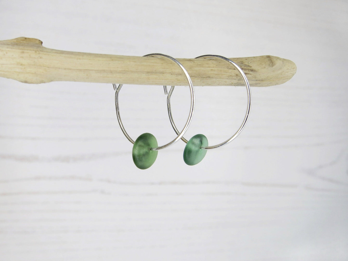 Silver hoop earrings with green sea glass hanging on a piece of driftwood against a white background