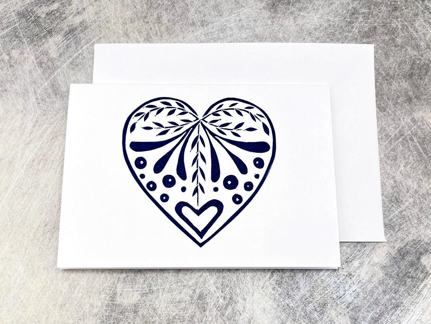 White card with an original lino printed  blue heart design stood on a textured surface. 