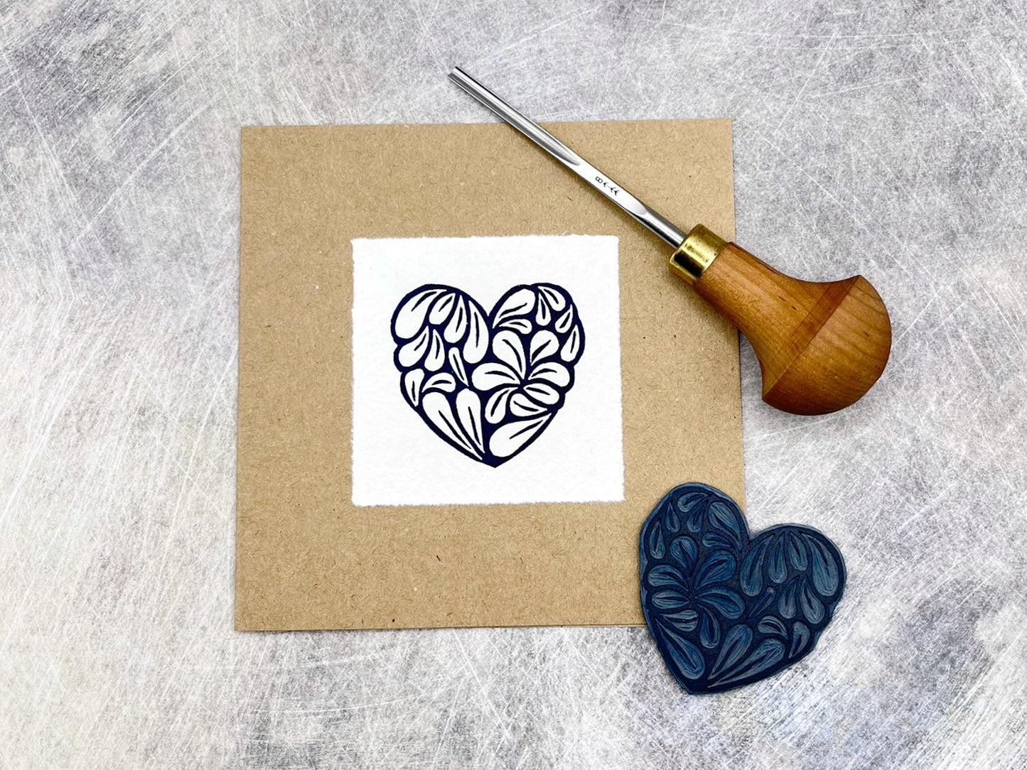 Kraft card with a white square featuring an original lino printed blue heart design, laid on a textured grey background. Photo includes lino and cutting tool used.