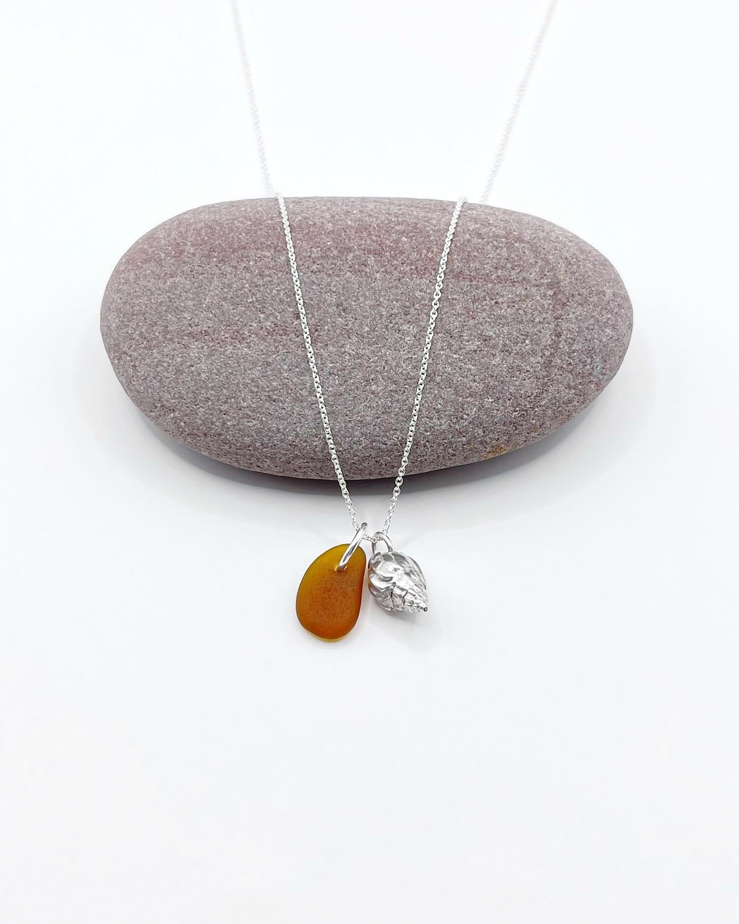 Necklace with burnt orange sea glass and silver shell pendant laid in front of a purple pebble on a white background