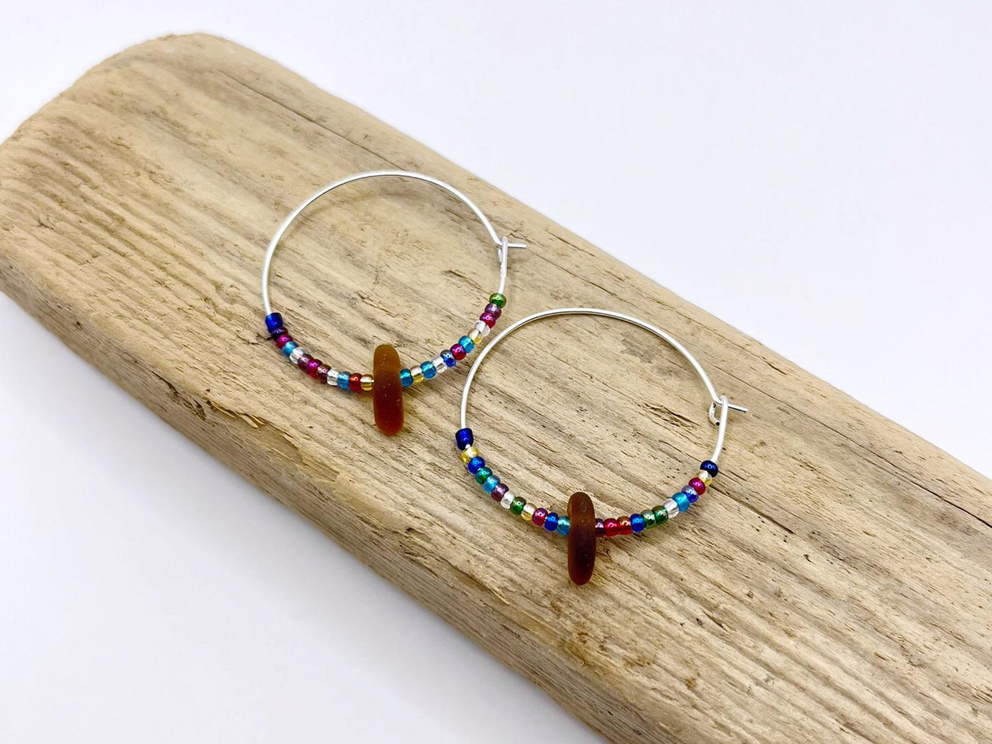 Rainbow seed bead and burnt orange sea glass hoop earrings laid on driftwood against a white background