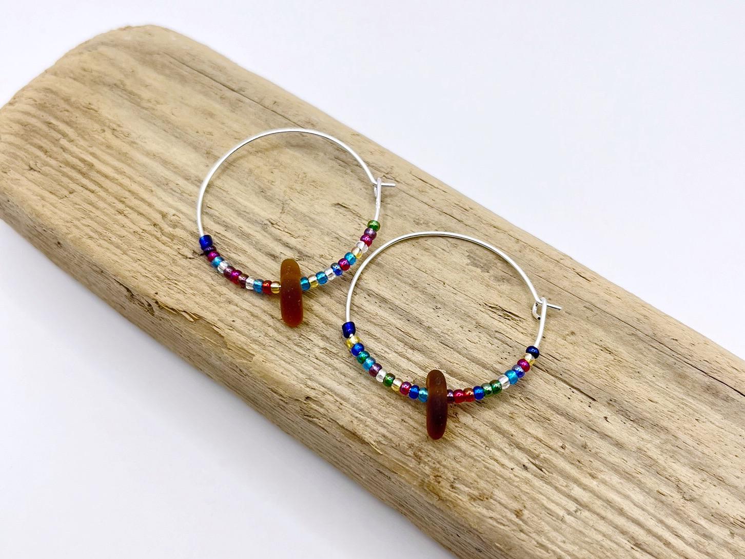 Rainbow seed bead and burnt orange sea glass hoop earrings laid on driftwood against a white background