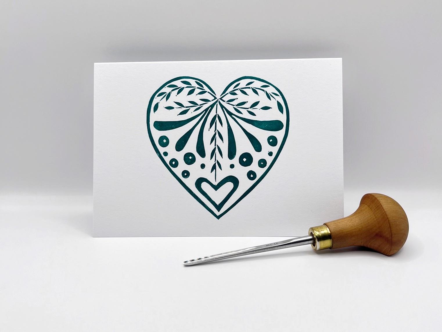 White card with an original lino printed green heart design stood on a white surface. Photo includes lino cutting tool.