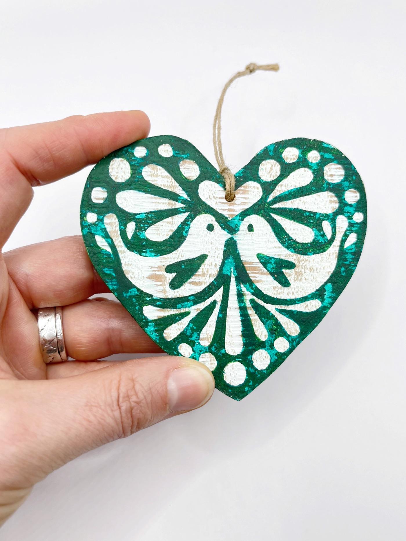 Green heart-shaped wooden hanging decoration with folk style love bird print   held in a hand against a white background