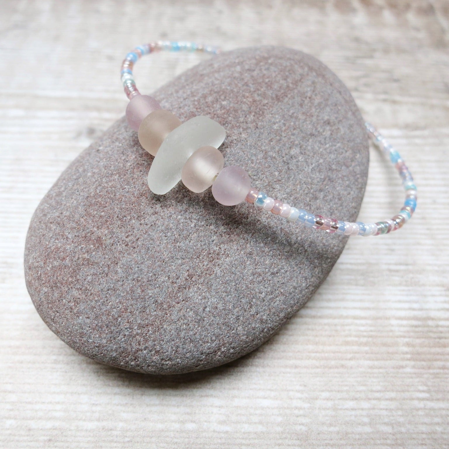 Bracelet with white sea glass and pastel beads laid on a purple pebble against a wooden background