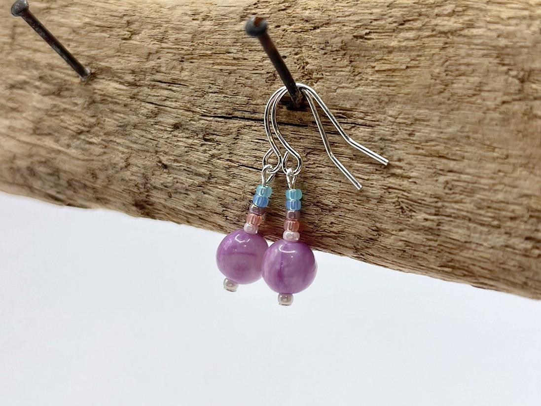 Pair of pastel seed bead and lilac jasper earrings hanging on driftwood against a white background.