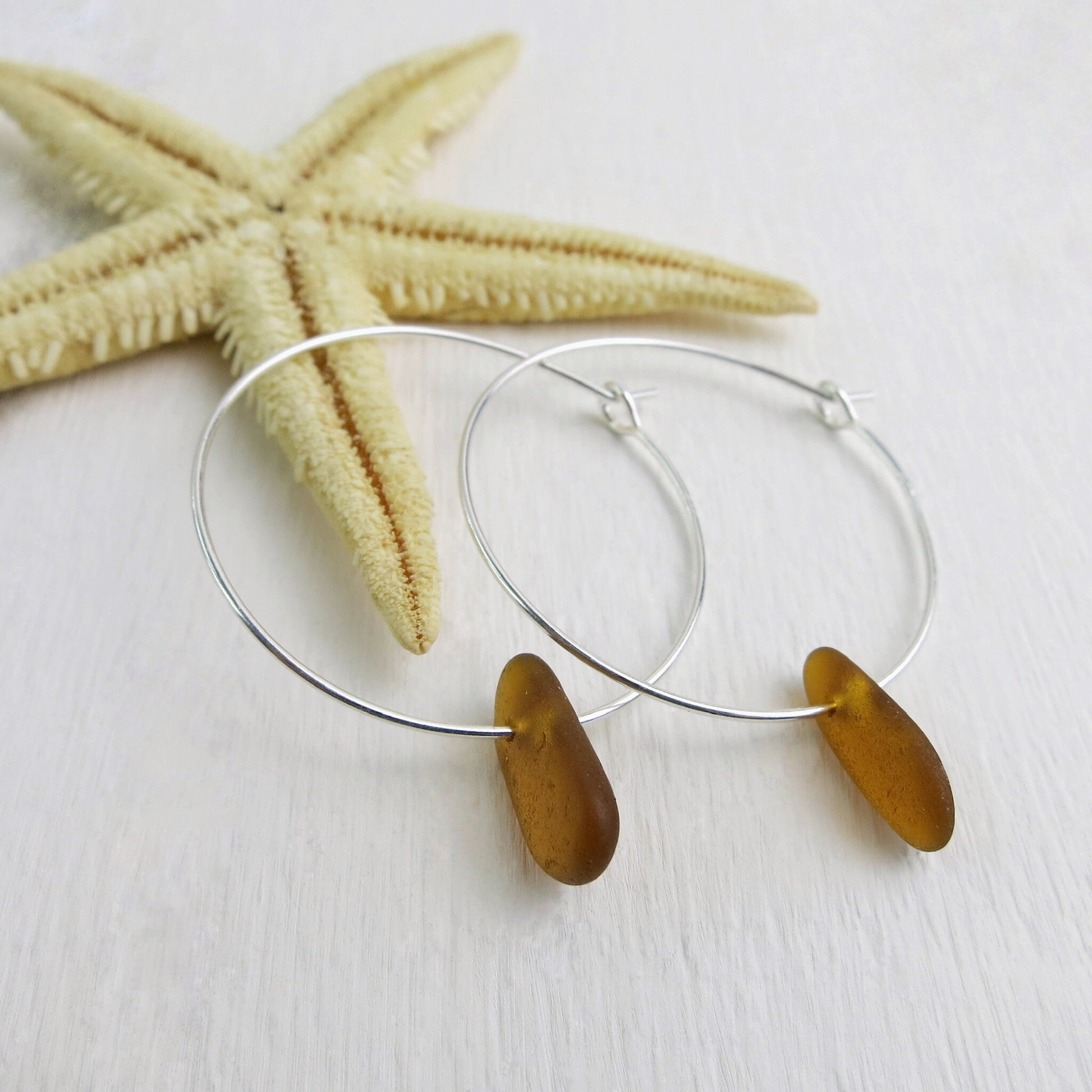 Silver hoop earrings with burnt orange sea glass laid against a starfish on a white surface.