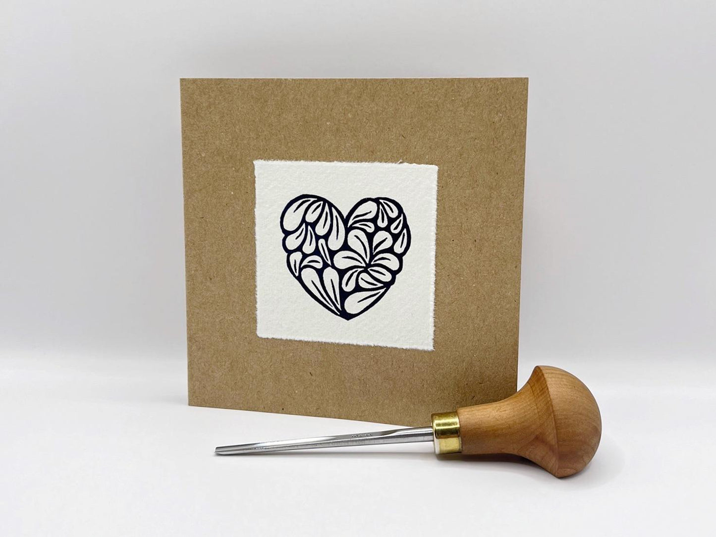 Kraft card with a white square featuring an original lino printed blue heart design, stood against a white background. Photo includes the lino cutting tool used.