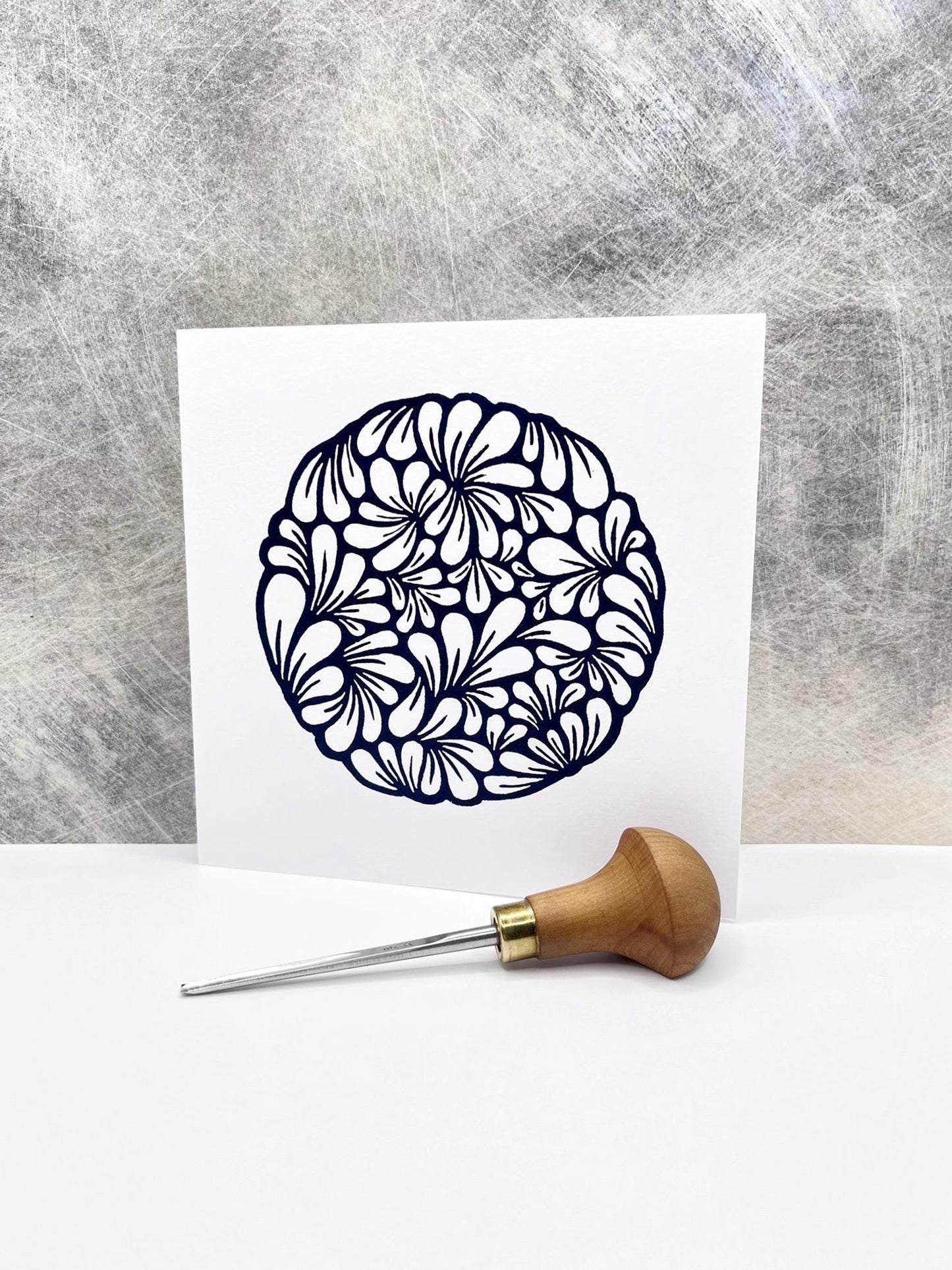 Original navy blue lino printed mussel shell pattern on white card stood against a textured background. Photo includes lino cutting tool.