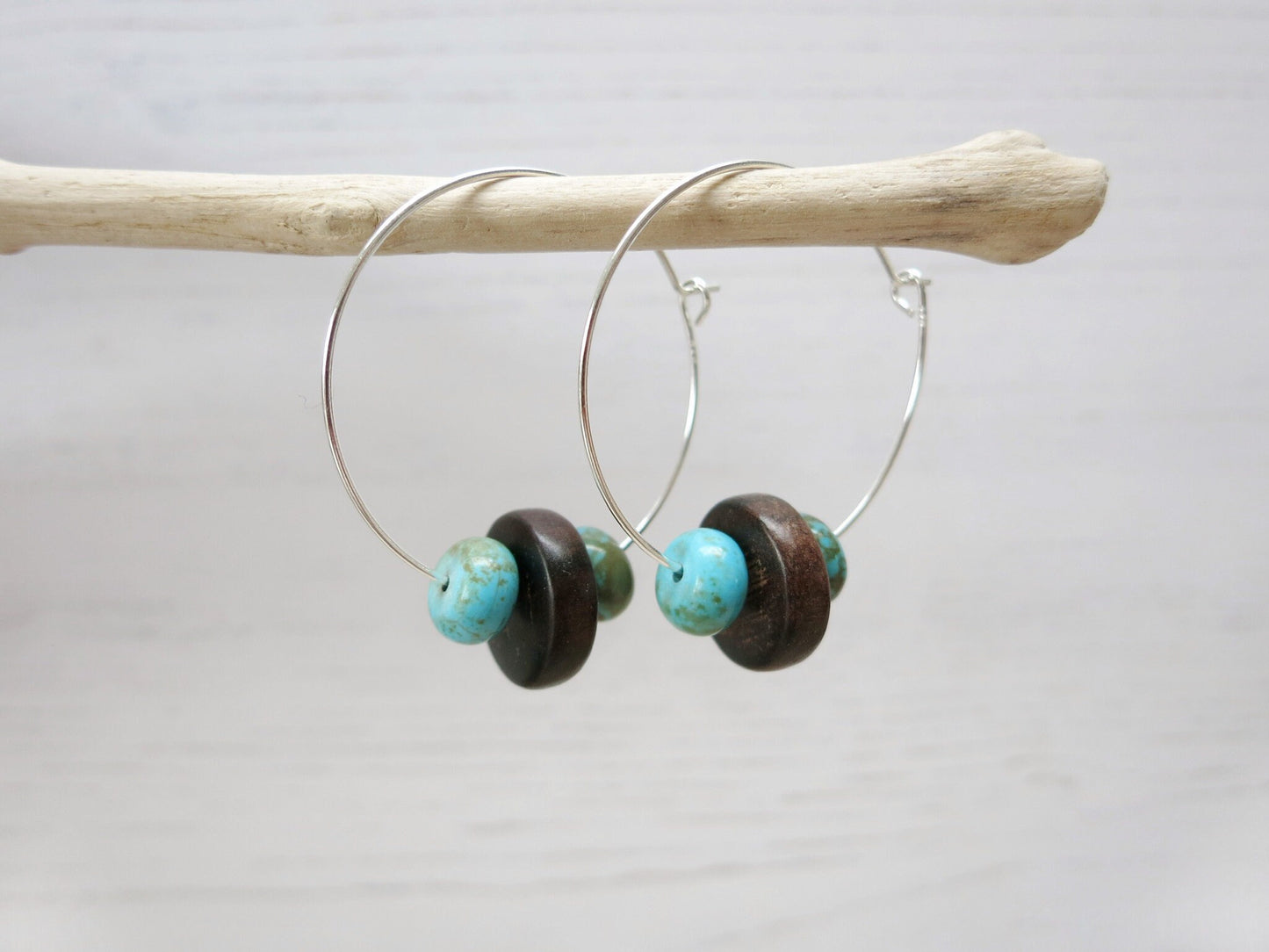 Close up of silver hoop earrings with turquoise and wooden beads hanging on driftwood against a light background