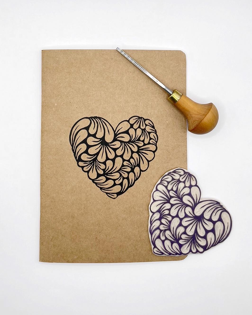 Kraft notebook with an original lino printed heart design laid on a white background. Photo includes lino and cutting tool used.