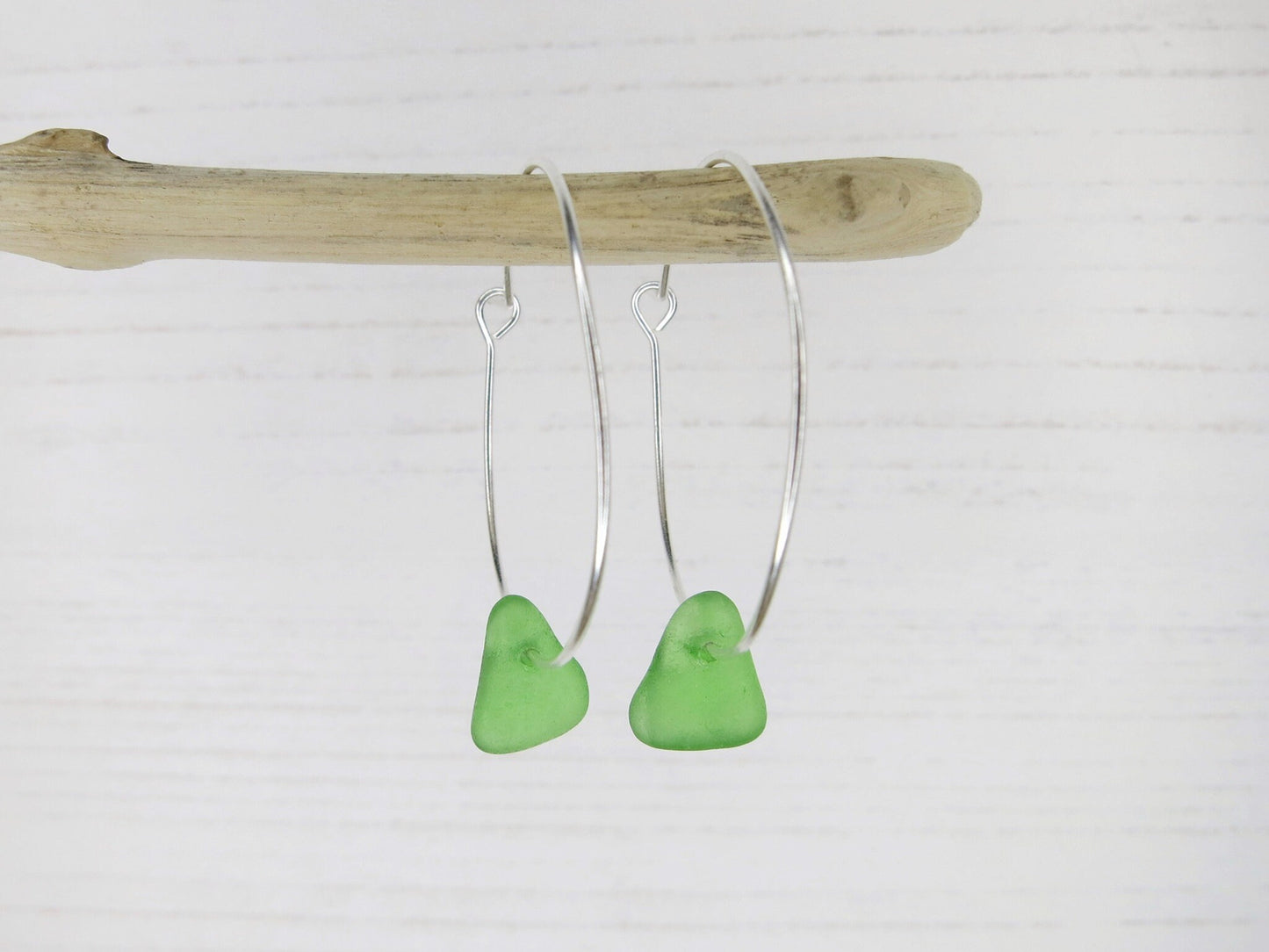 Green sea glass silver hoop earrings hanging on a piece of driftwood against a white background