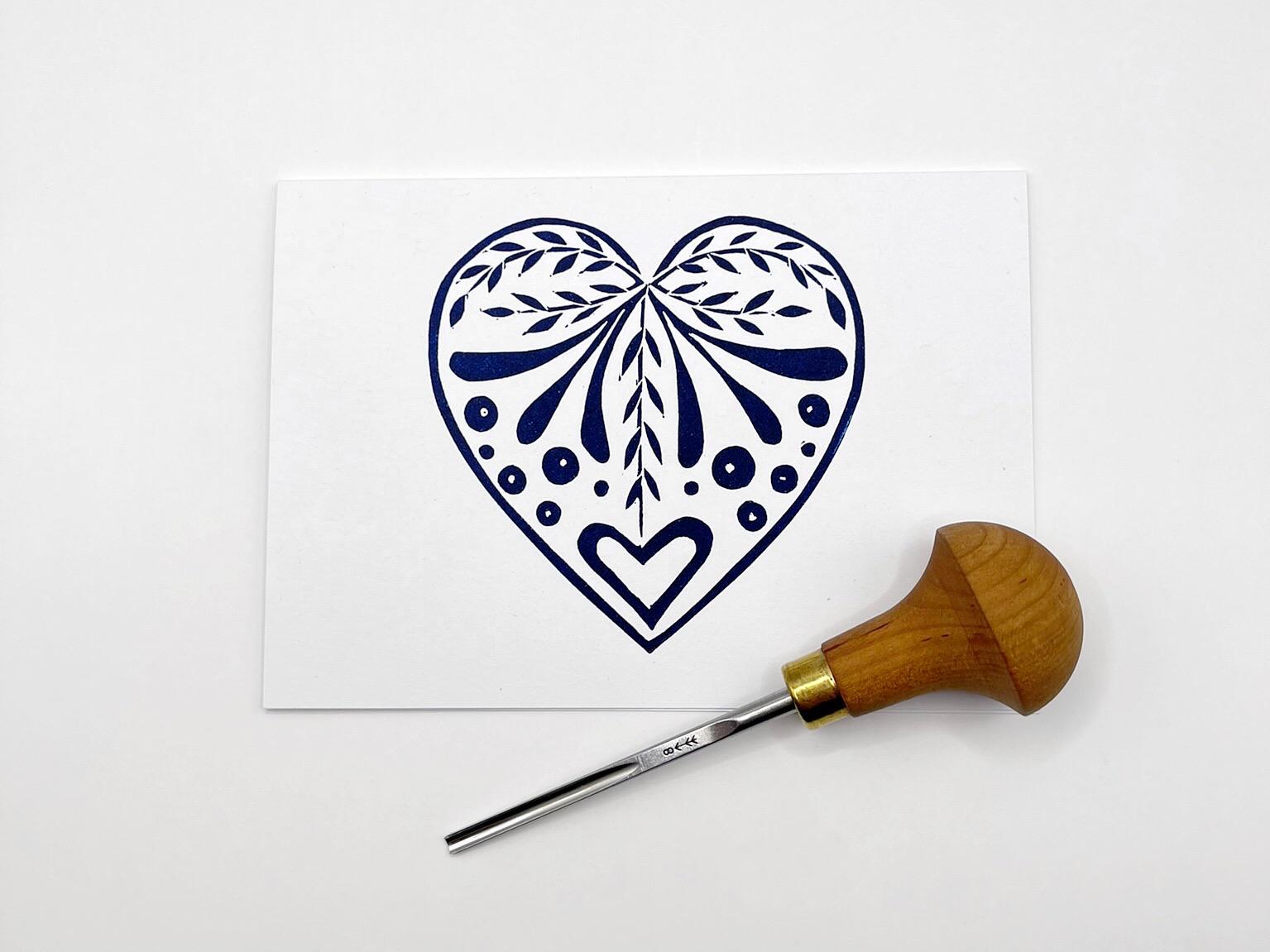 White card with an original lino printed  blue heart design laid on a white surface. Photo includes lino cutting tool.