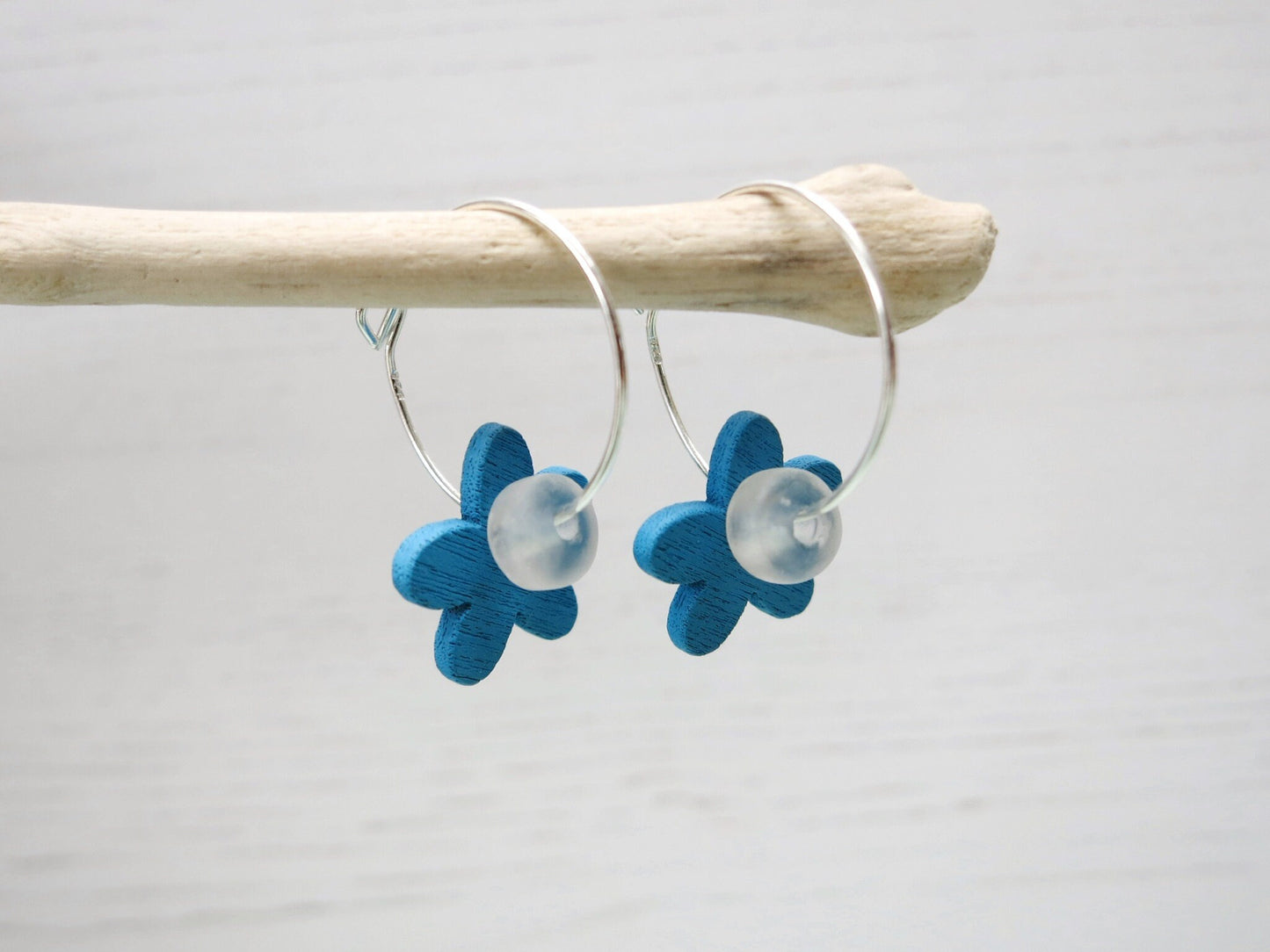 Side view of silver hoop earrings with Blue wooden flowers and a glass bead hanging on driftwood against a white background