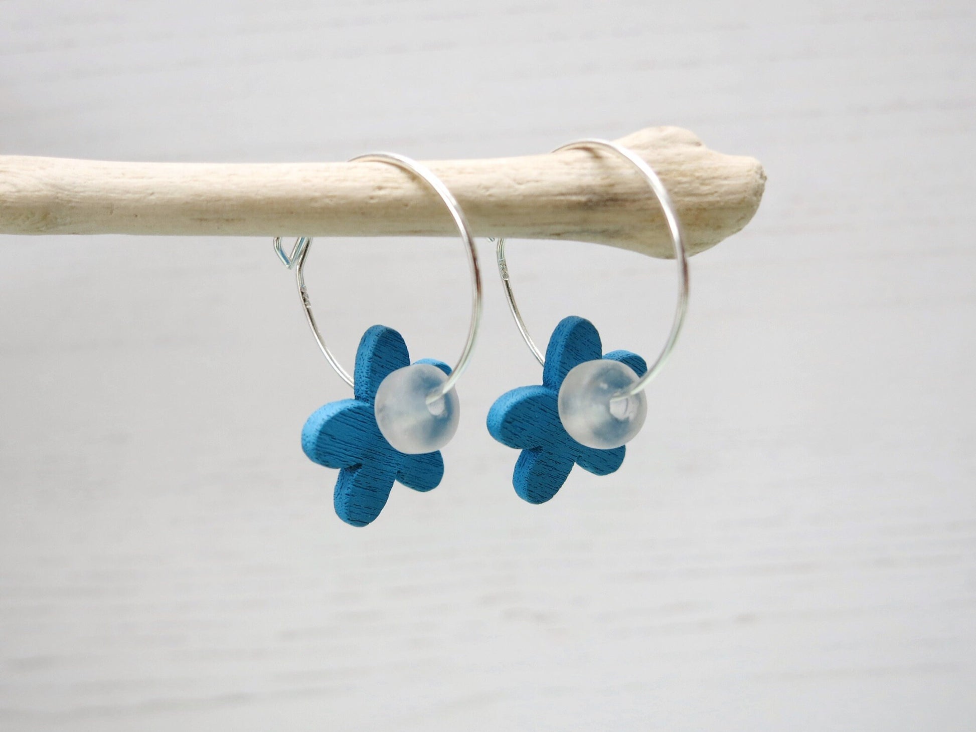 Side view of silver hoop earrings with Blue wooden flowers and a glass bead hanging on driftwood against a white background