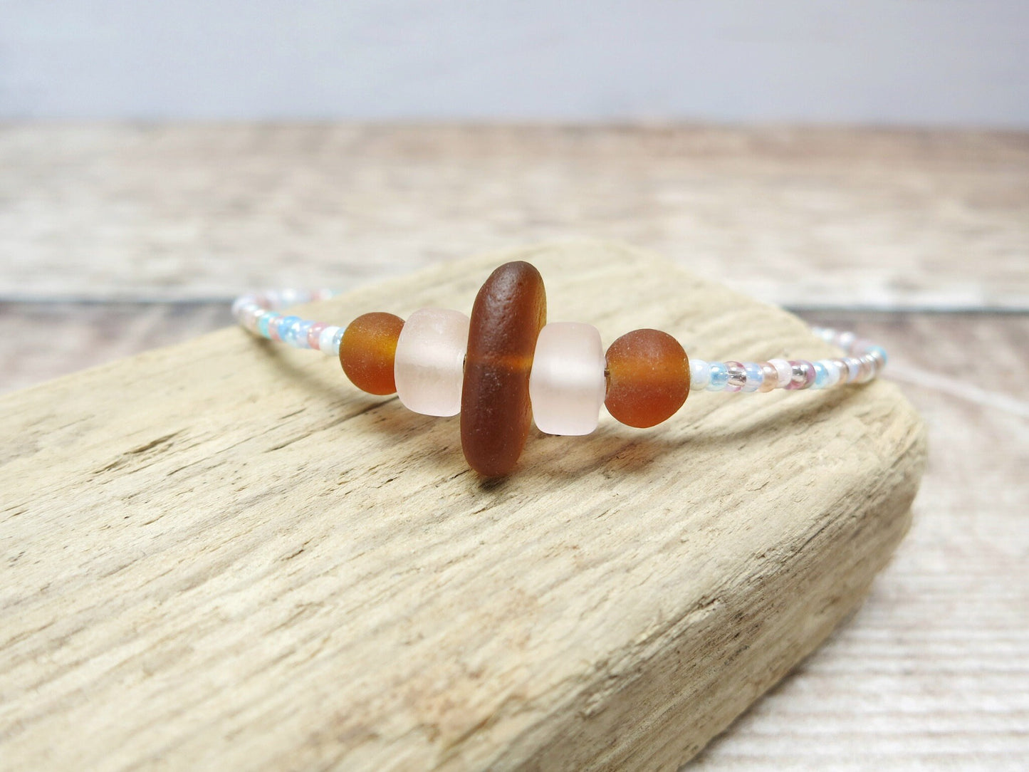 Close up of bracelet with pastel seed beads and burnt orange sea glass laid on driftwood.