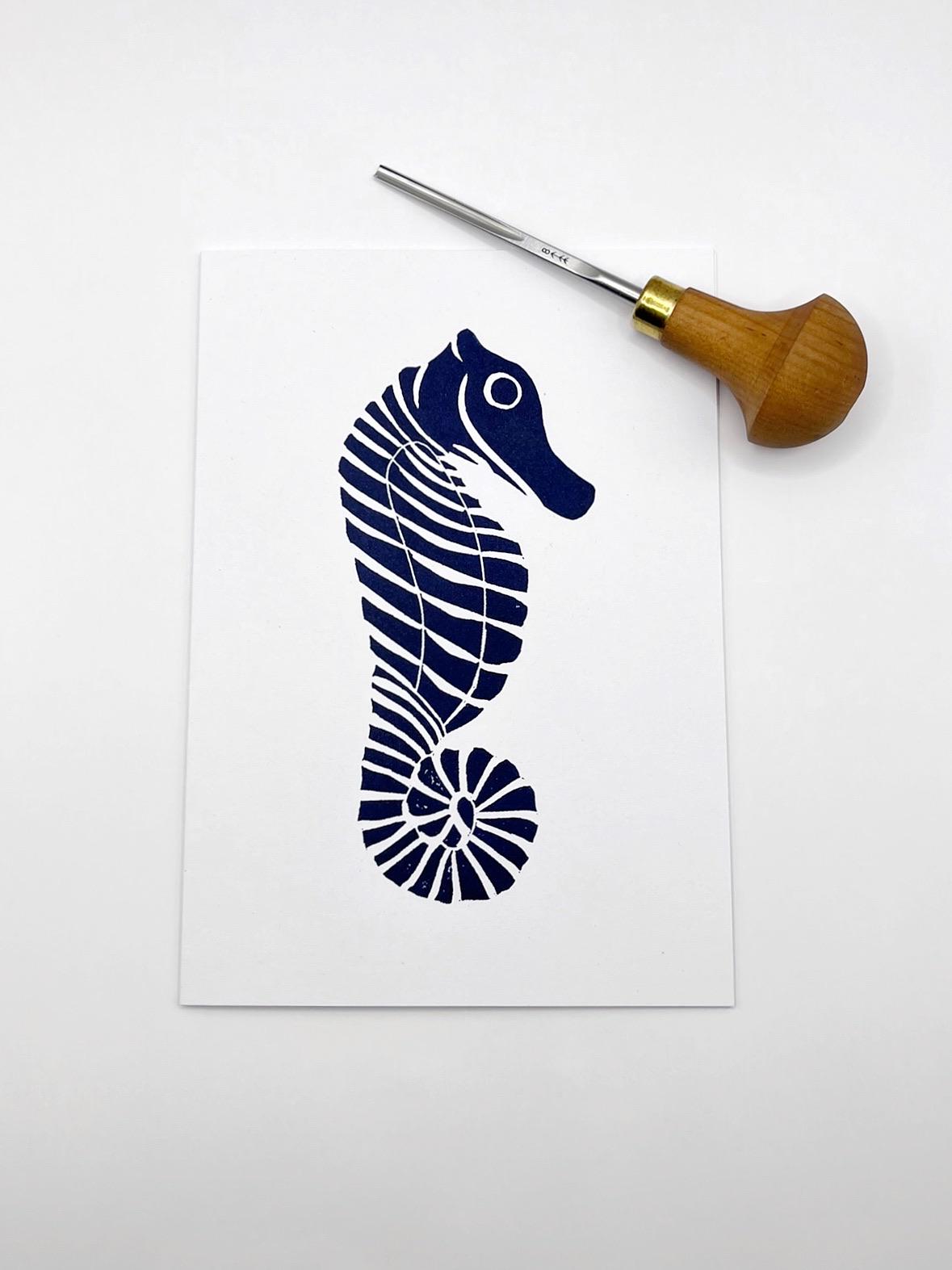 Original lino print of a navy blue seahorse design on a white card, laid on a white background. Photo includes lino cutting tool.
