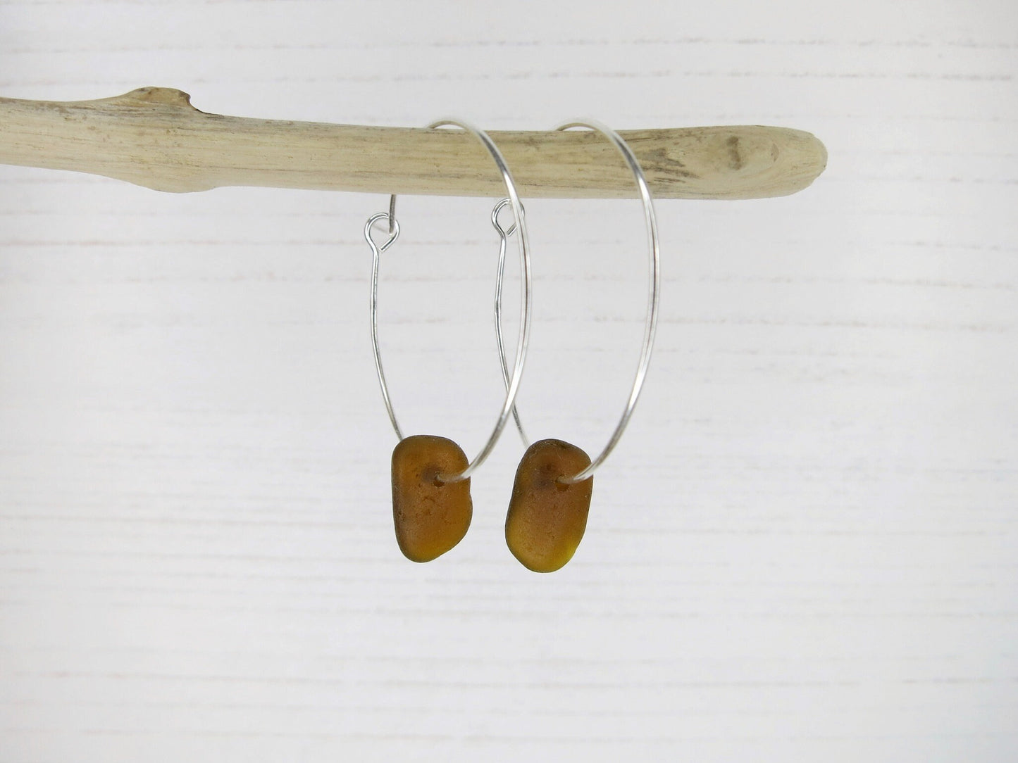 Silver hoop earrings with burnt orange sea  glass hanging on driftwood against a white background