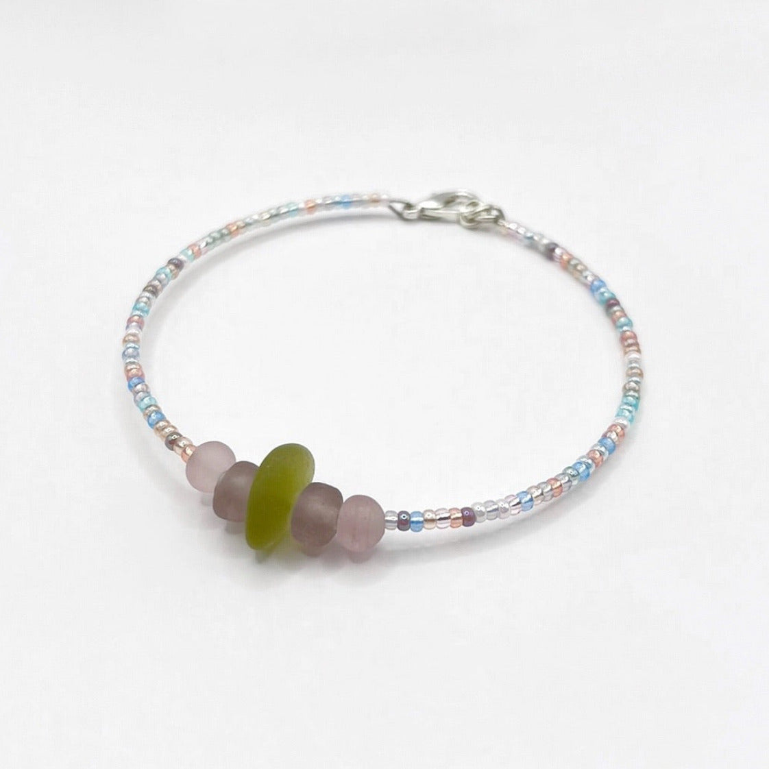 A pastel beaded bracelet with purple and lilac frosted glass beads and a piece of soft lime coloured Cornish sea glass, finished with a silver plated lobster clasp and laid on a white background.
