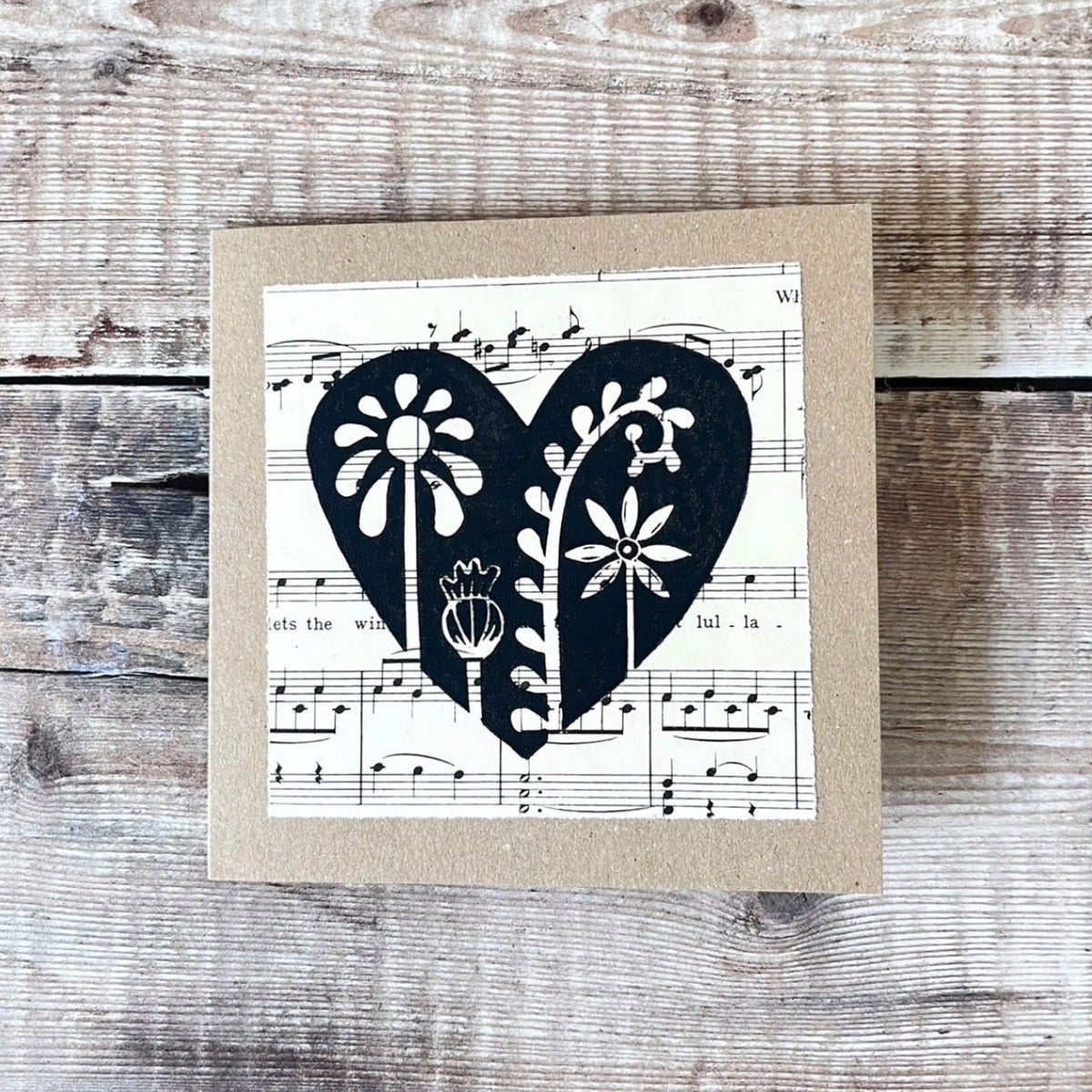 Square kraft greeting card with a black floral heart lino print on vintage music paper mounted on the front. Laid on a wooden background.