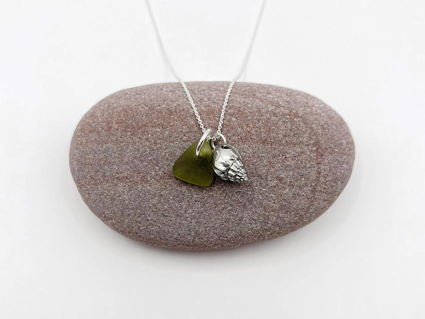 Necklace with olive green sea glass and a silver shell charm laid on a pebble against a white background