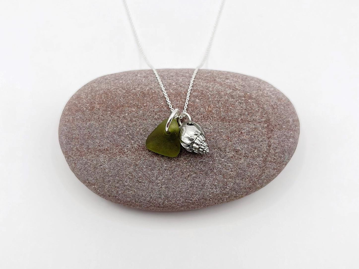 Necklace with olive green sea glass and a silver shell charm laid on a pebble against a white background