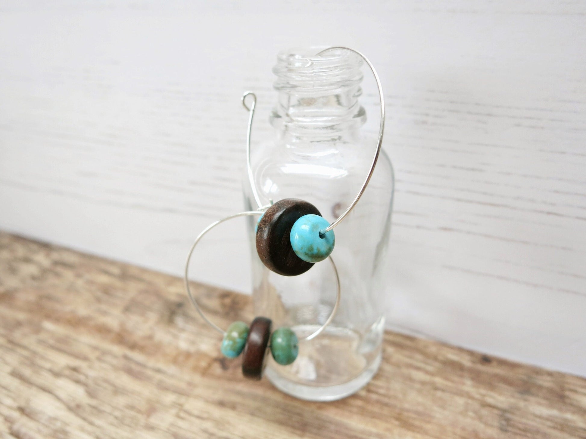 Close up of silver hoop earrings with turquoise and wooden beads hanging on a glass bottle against a light background