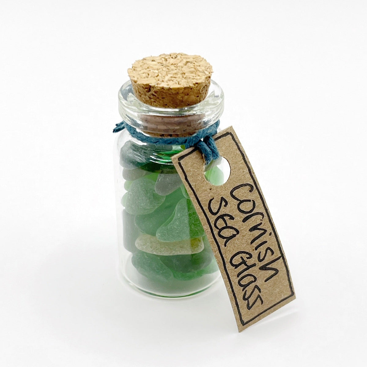 A small clear glass bottle with a cork stopper, filled with green sea glass pieces and tied with a blue string. A hanging kraft paper tag reads 'Cornish Sea Glass' on a clean white background.