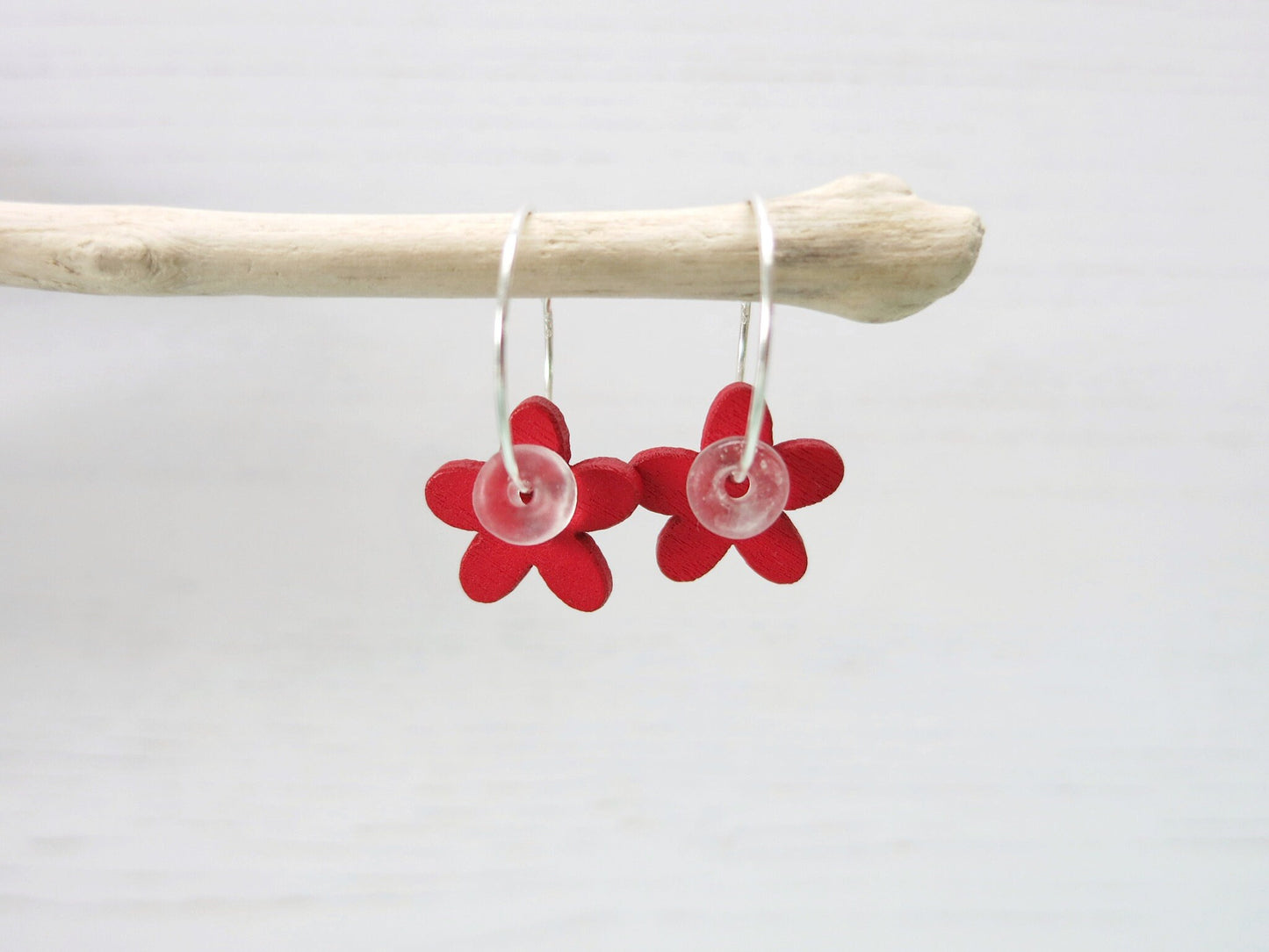 Silver hoop earrings with red wooden flowers and a glass bead hanging on driftwood against a white background