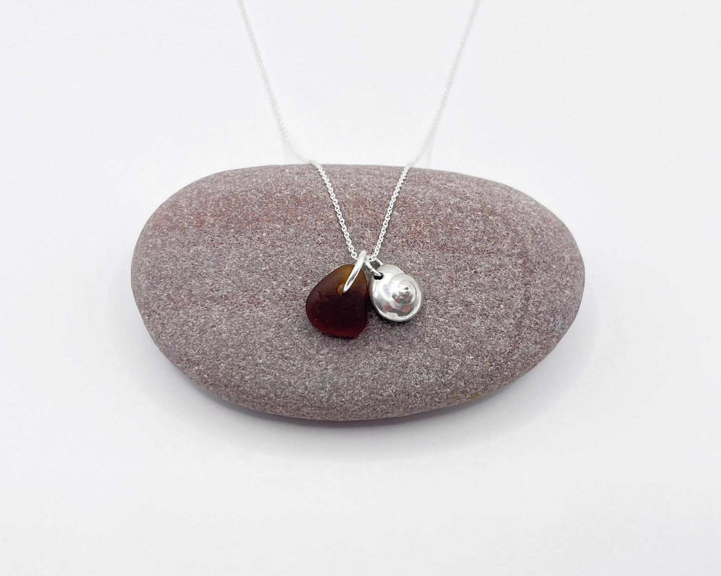 Necklace with burnt orange sea glass and a silver shell pendant, laid across a purple pebble on a white surface