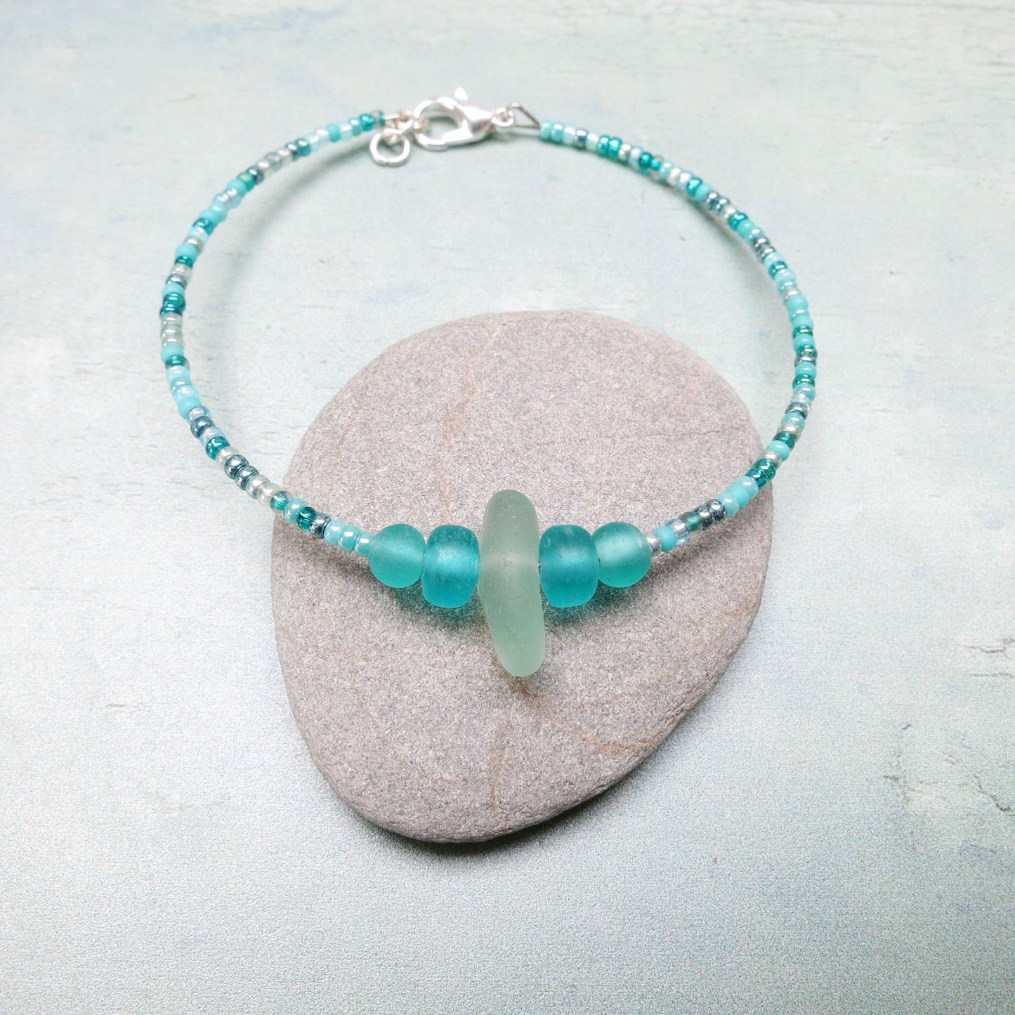Bracelet with pale sea foam sea glass and sea green and turquoise glass beads laid on a grey pebble against a pale blue background
