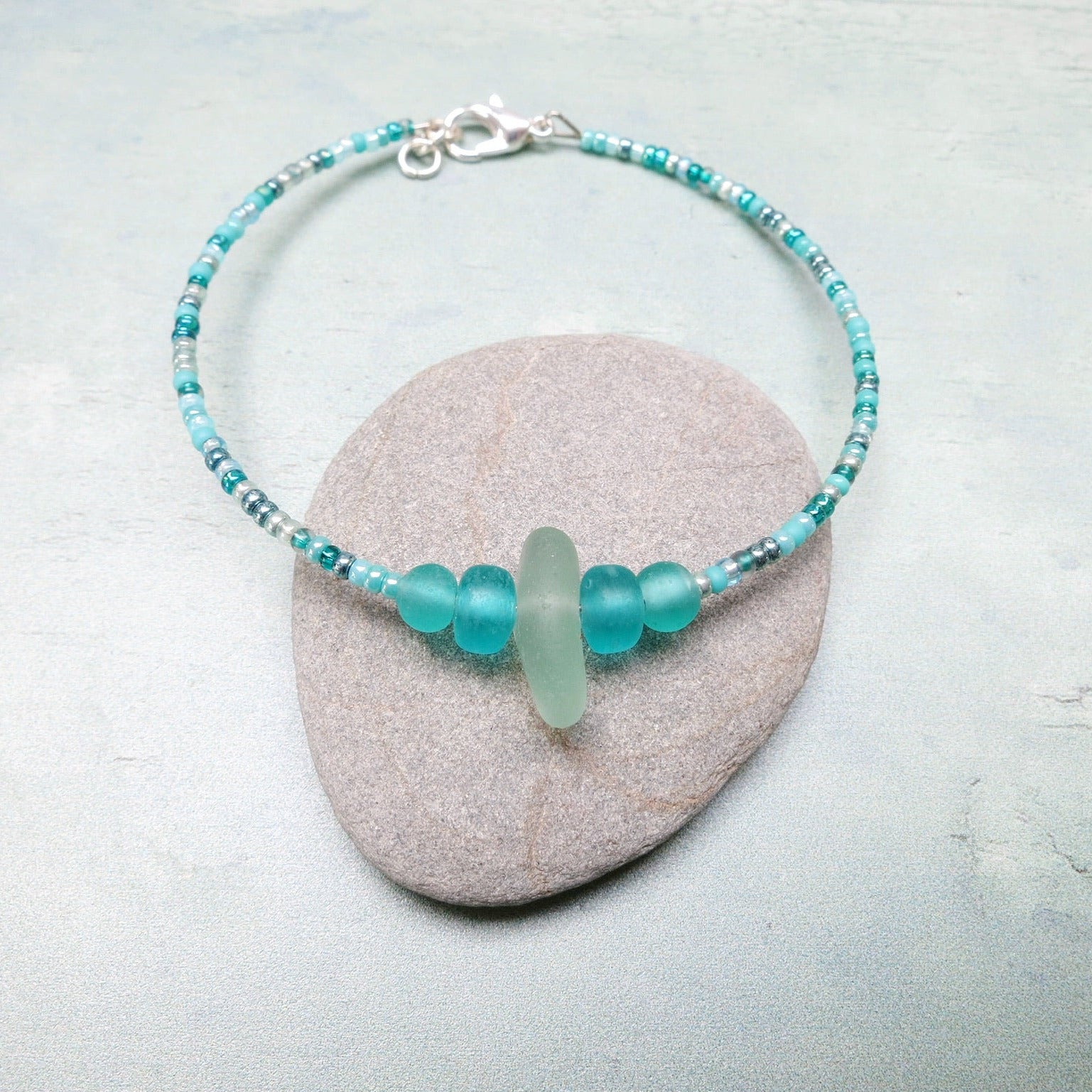 Bracelet with pale sea foam sea glass and sea green and turquoise glass beads laid on a grey pebble against a pale blue background
