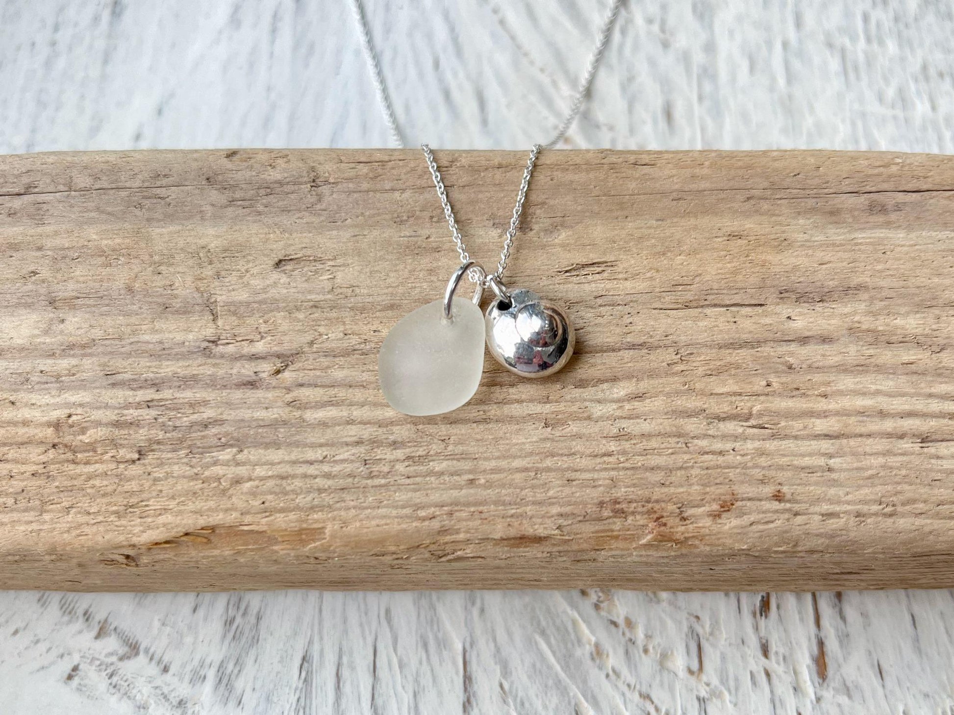 Necklace with a silver shell and white sea glass pendant laid on driftwood against a textured surface