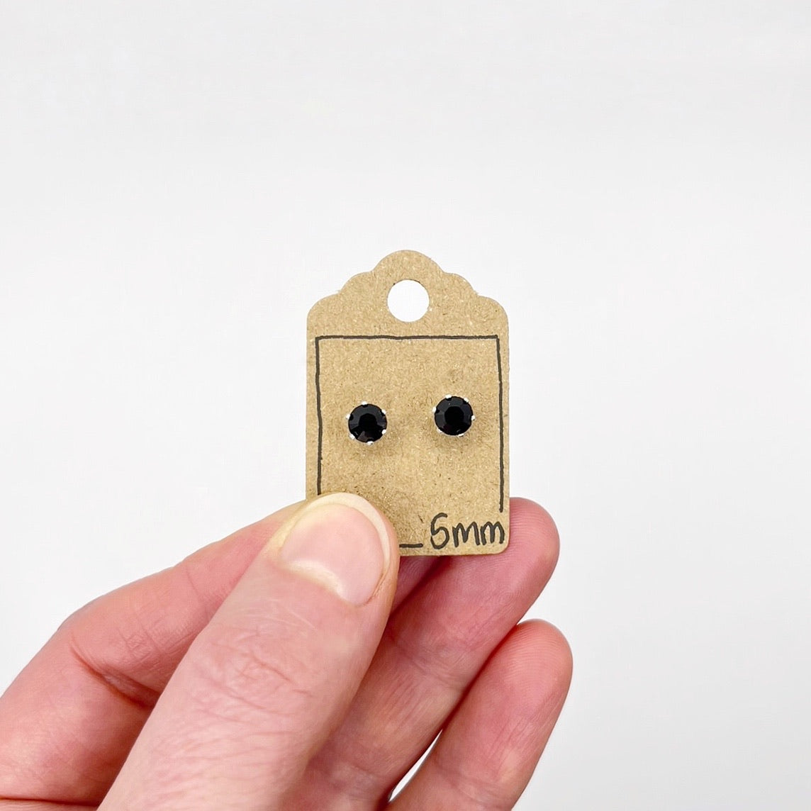 Small black stud earrings on a cardboard tag held by a hand against a white background